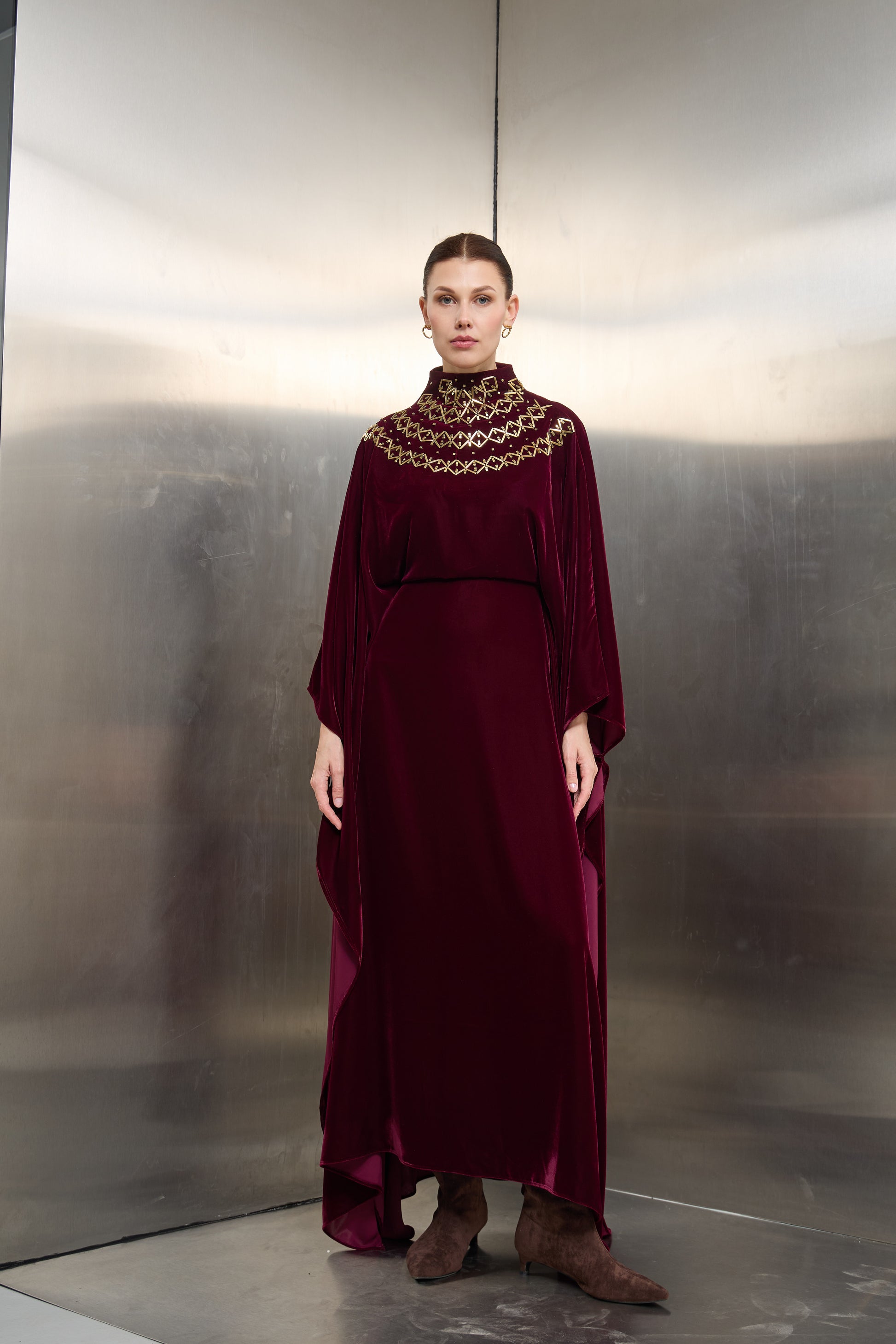 "Mona Al Shebil Burgundy velvet and crepe kaftan featuring a floor-length cape silhouette and a gold-stone embellished mock neck for luxury Saudi modest wear."