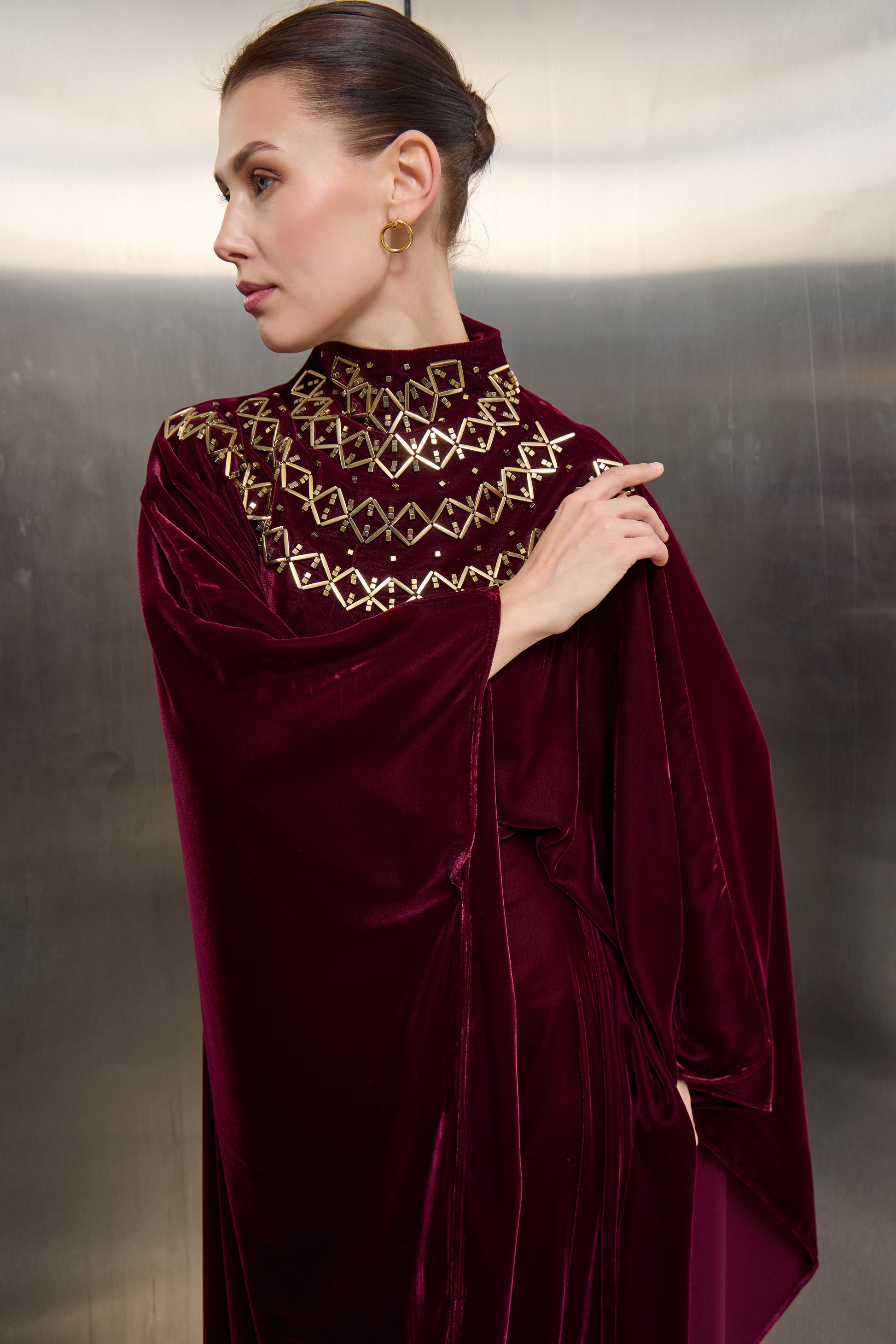 Detailed close-up of gold geometric stone embellishments on the burgundy velvet mock neck of a Mona Al Shebil luxury kaftan, highlighting high-end craftsmanship