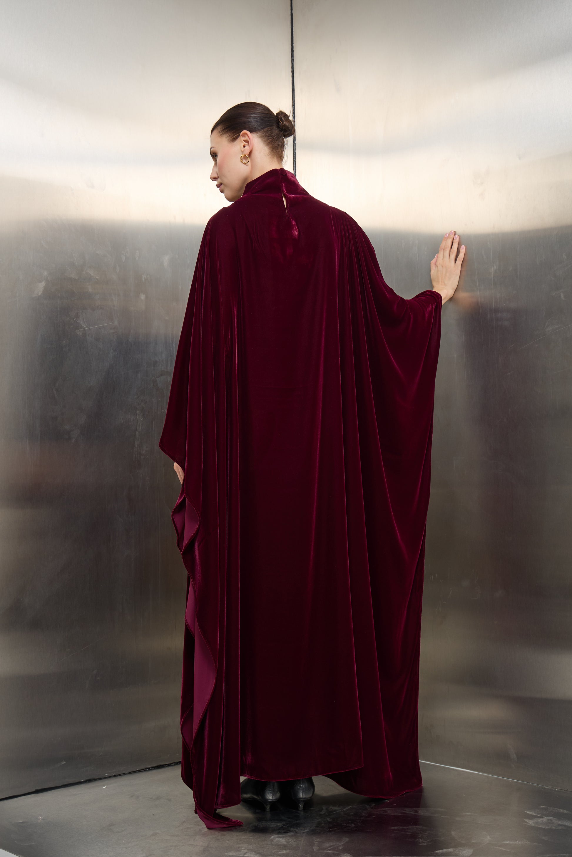 Back view of the burgundy velvet cape kaftan by Mona Al Shebil, showcasing the elegant floor-length drape and structured professional silhouette.