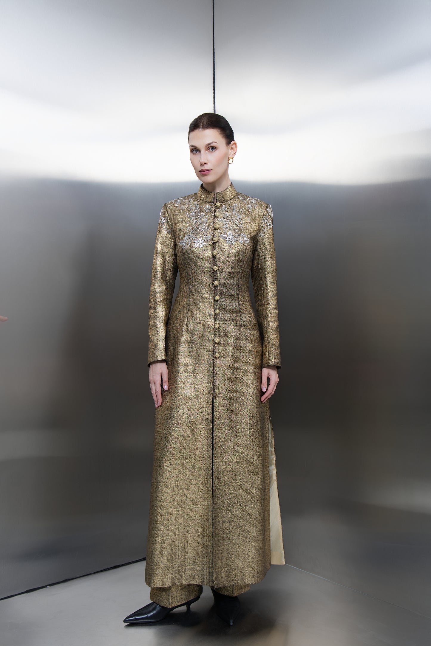 "A full-length view of the Mona Al Shebil Embellished Gold Structured Long Jacket, showcasing a regal metallic gold weave, mandarin collar, and center-front button detailing paired with matching wide-leg trousers for a high-end Saudi luxury aesthetic."